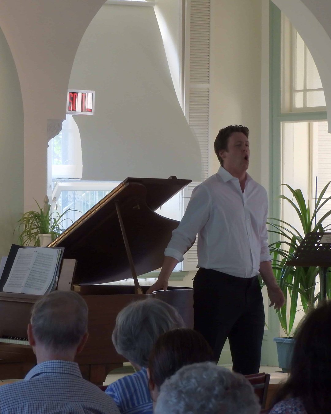 Un chanteur, accompagné au piano, devant le public attentif d'une salle aux arches blanches.