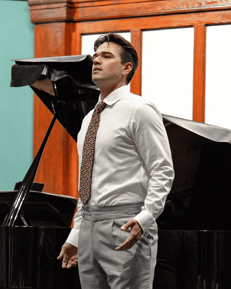 Un chanteur en chemise blanche et cravate, debout devant un piano à queue.