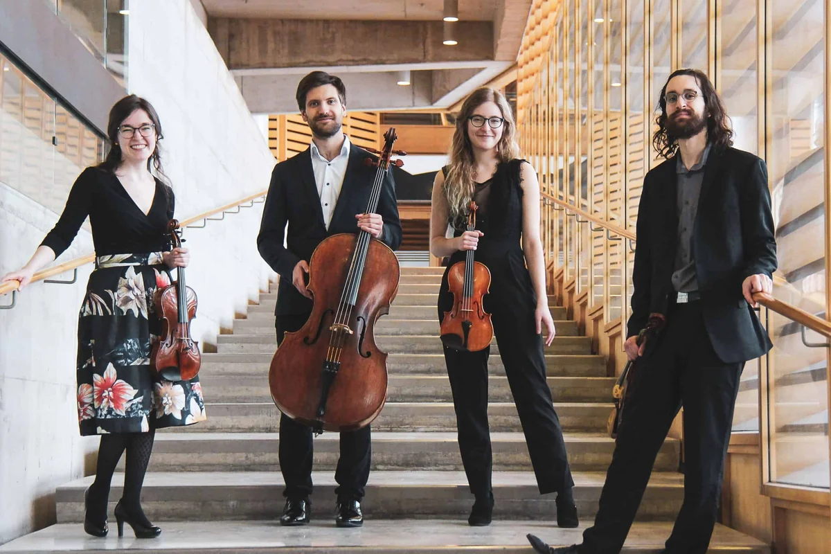 Quatre musiciens à cordes posent avec leurs instruments dans un escalier de bois clair.